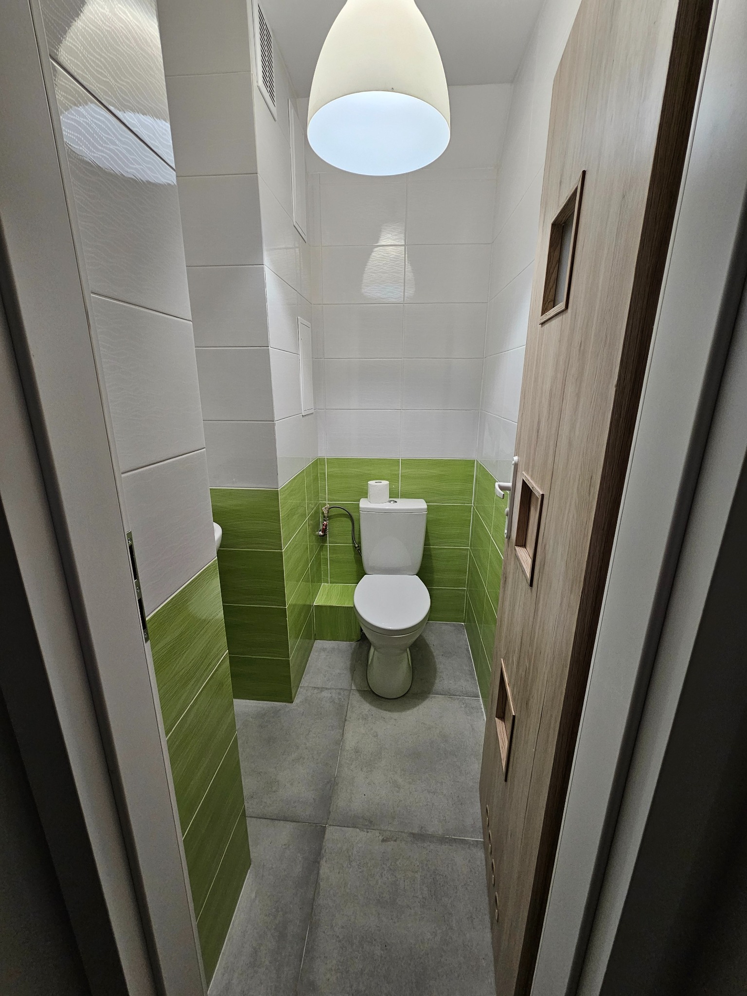 Wnętrze łazienki z toaletą, wykończone białymi i zielonymi płytkami, szarą podłogą oraz drewnianymi drzwiami z minimalistycznymi przeszkleniami.
