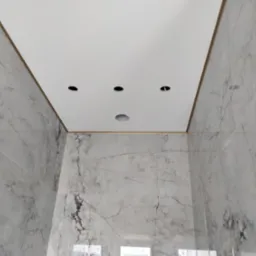Widok w górę na sufit podwieszany z czterema otworami na oświetlenie punktowe, otoczony marmurowymi płytkami ściennymi, w trakcie instalacji oświetlenia.