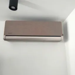 Szaro-beżowy klimatyzator marki Rotenso zamontowany na białej ścianie w pomieszczeniu, widoczna lampa sufitowa.