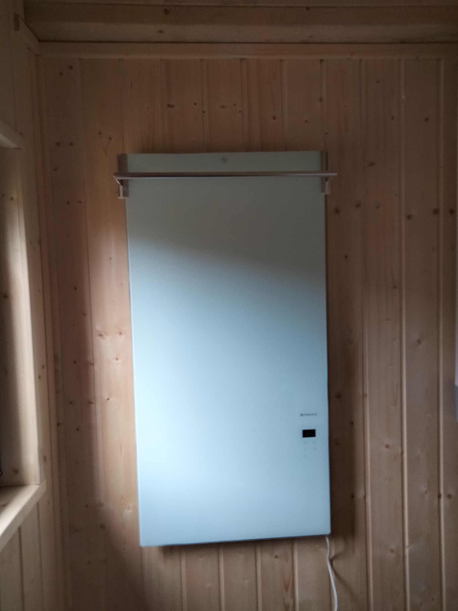 Biały panel grzewczy zawieszony na drewnianej ścianie z boazerii, widoczny kabel zasilający i metalowy uchwyt nad panelem.