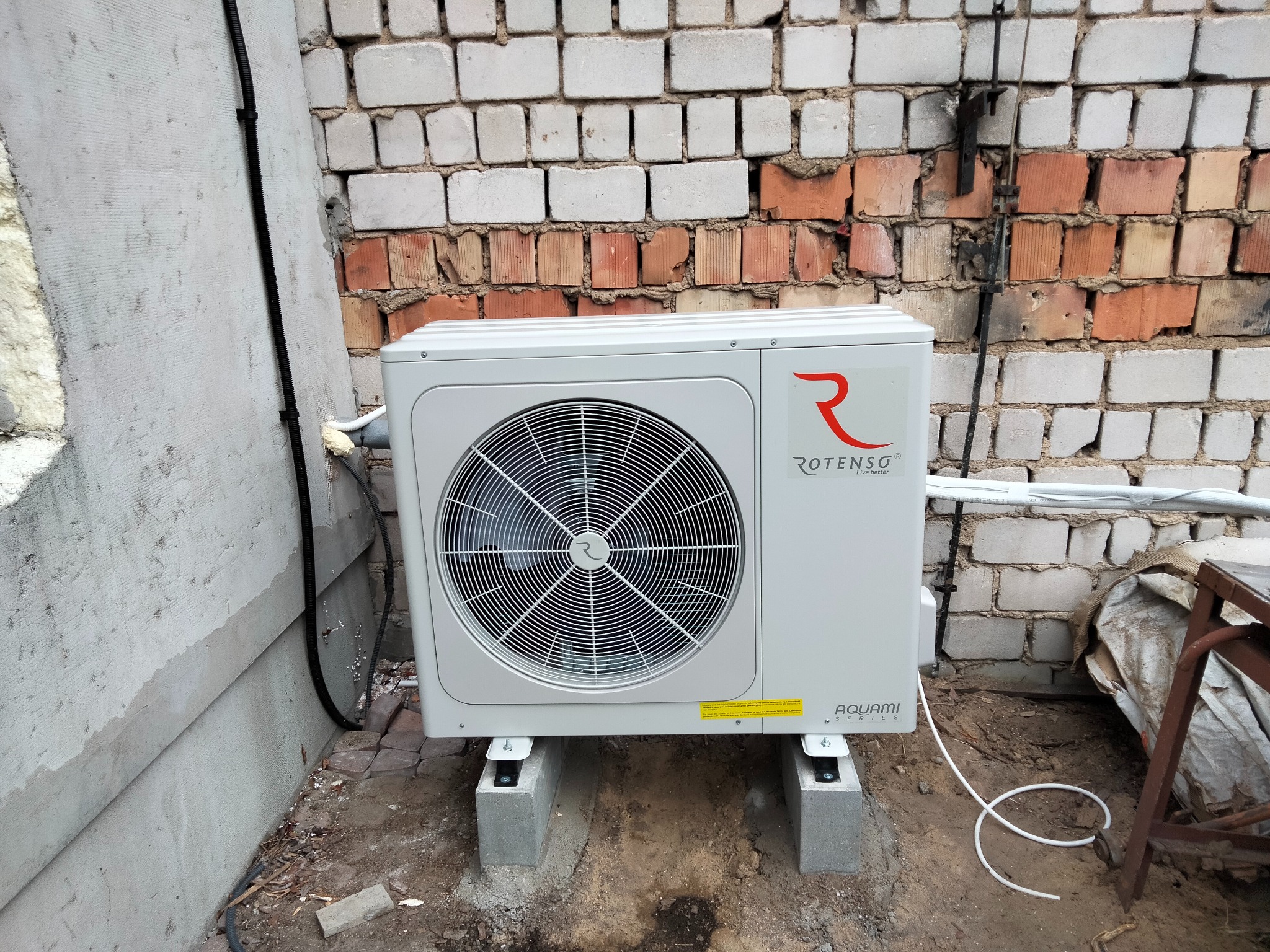 Biała jednostka zewnętrzna pompy ciepła marki Rotenso Aquami Series, zamontowana na betonowych bloczkach przy ceglanym murze z widocznymi przewodami i ziemią.