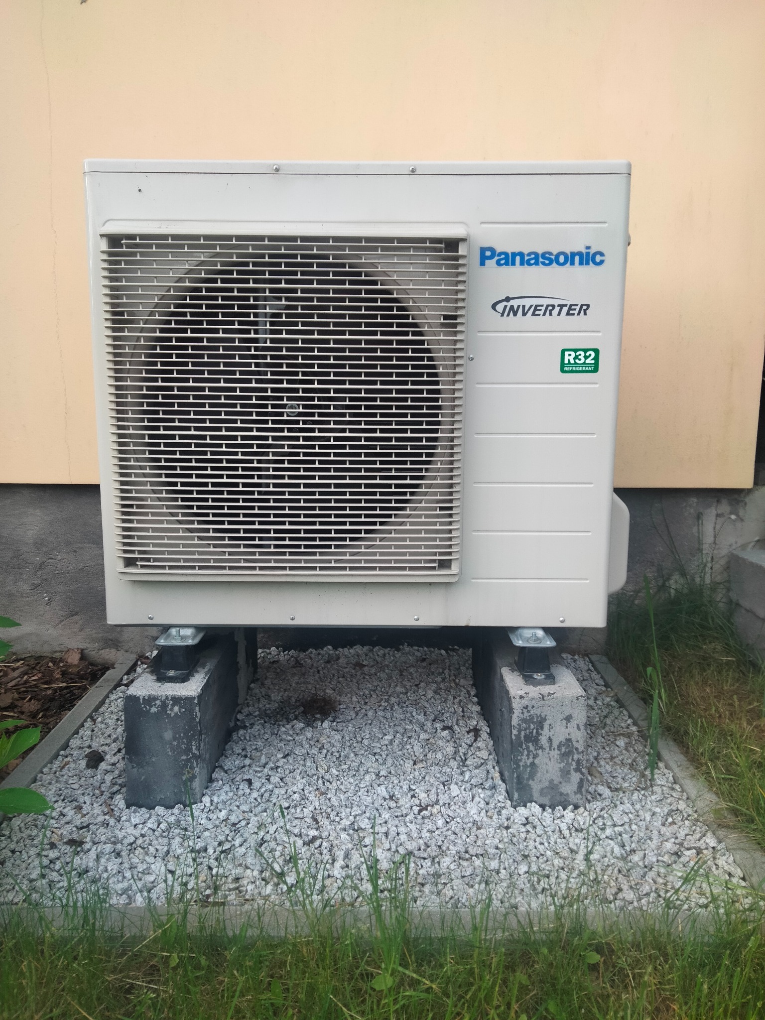 Zewnętrzna jednostka klimatyzacji Panasonic Inverter R32, ustawiona na betonowych blokach na żwirowym podłożu, z fragmentem elewacji budynku w tle.