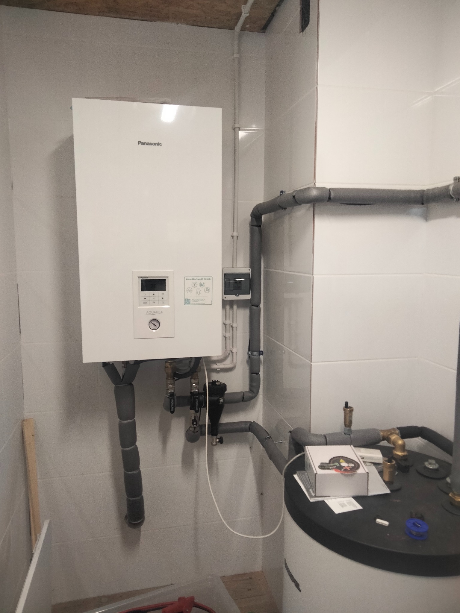 Biała pompa ciepła Panasonic Aquarea, zamontowana na ścianie z białych płytek, z widocznymi szarymi rurami izolowanymi i zbiornikiem na wodę, w pomieszczeniu o surowym wykończeniu.