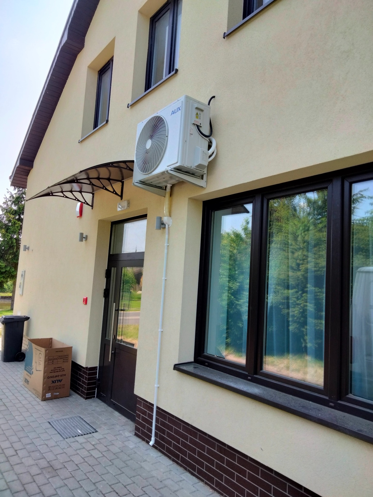 Zewnętrzna jednostka klimatyzacji AUX zamontowana na elewacji budynku z beżową fasadą, widoczny przewód odprowadzający skropliny, okno z ciemną ramą i wejście do budynku z daszkiem.