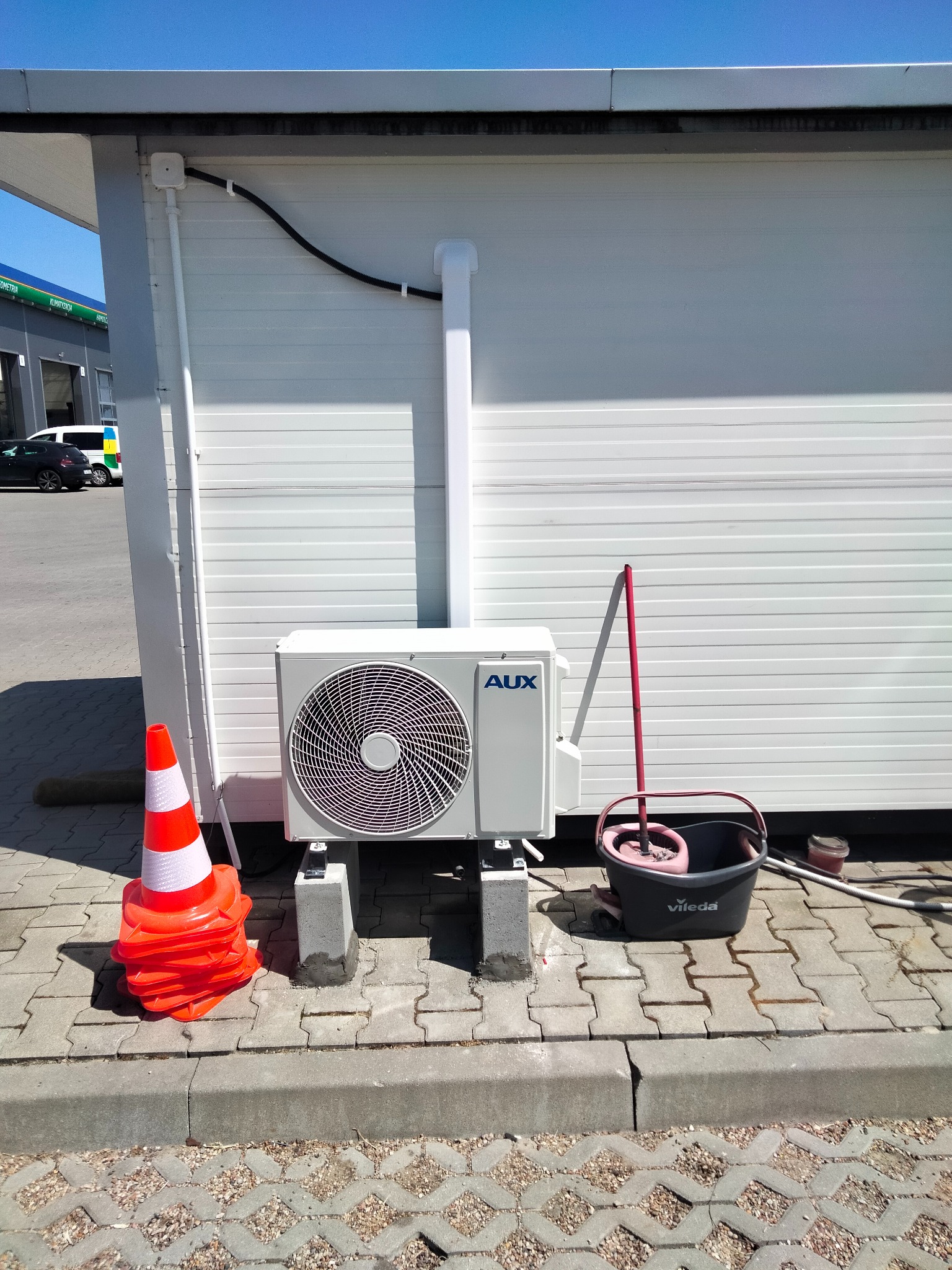 Zewnętrzna jednostka klimatyzacji AUX zamontowana na betonowych wspornikach przy białej ścianie budynku, obok leżą ułożone pomarańczowe pachołki drogowe i stoi wiadro z mopem Vileda.