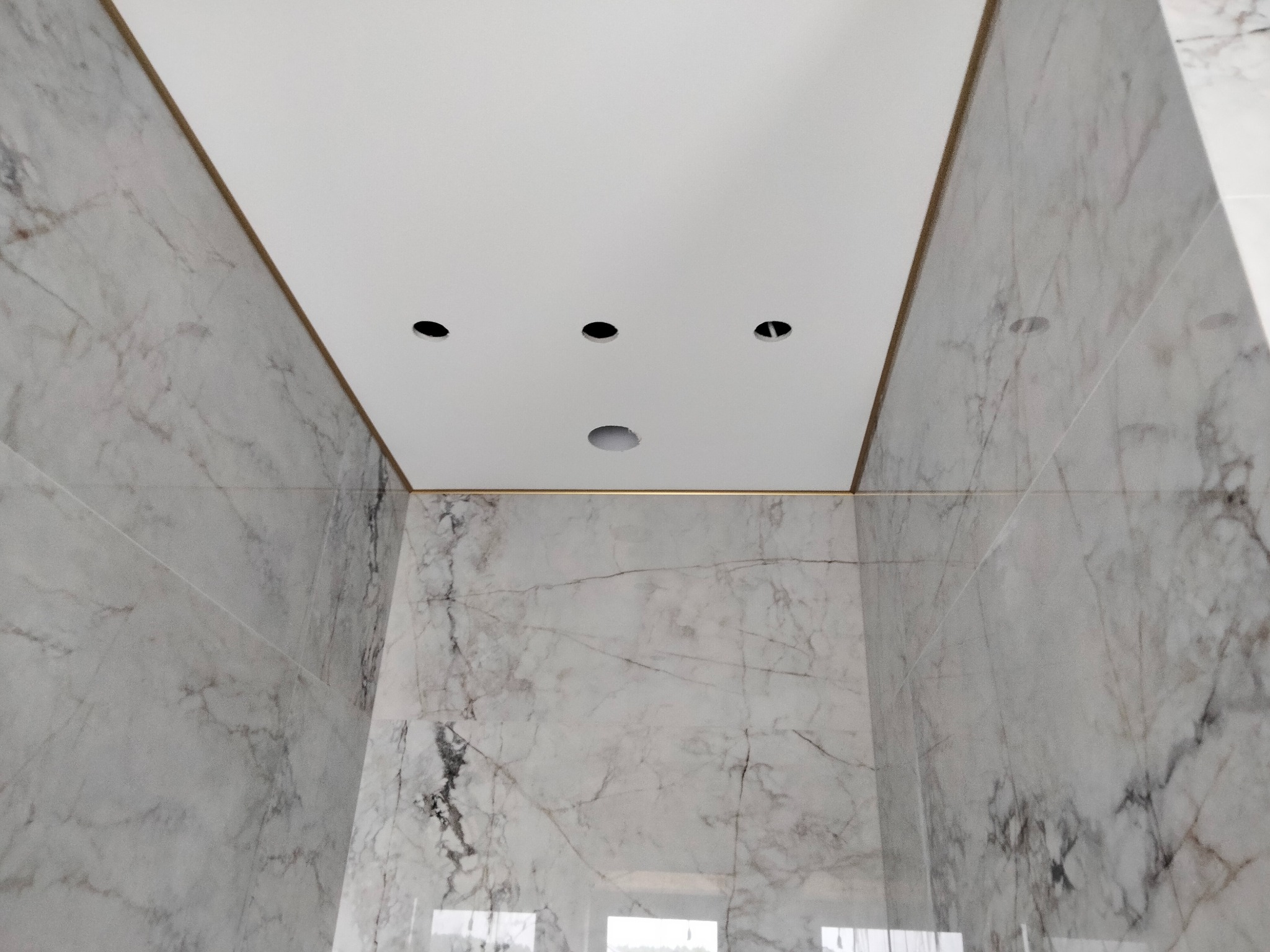 Widok w górę na sufit podwieszany z czterema otworami na oświetlenie punktowe, otoczony marmurowymi płytkami ściennymi, w trakcie instalacji oświetlenia.