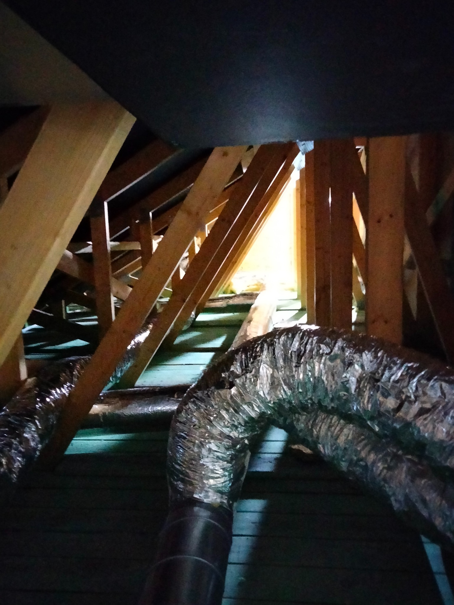 Montaż elastycznych przewodów wentylacyjnych w przestrzeni poddasza, widoczne drewniane elementy konstrukcyjne dachu i szczelina z widokiem na światło dzienne.