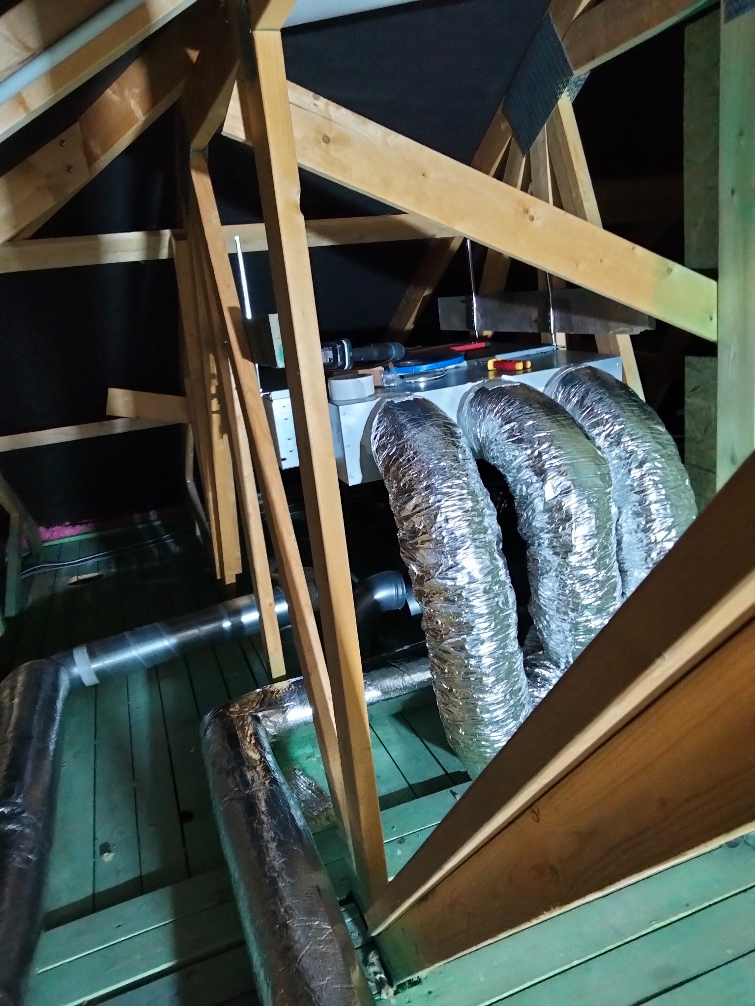 Instalacja rekuperacji na poddaszu: srebrne, elastyczne przewody wentylacyjne podłączone do centrali, widoczne drewniane elementy konstrukcji dachu i zielona podłoga z desek.