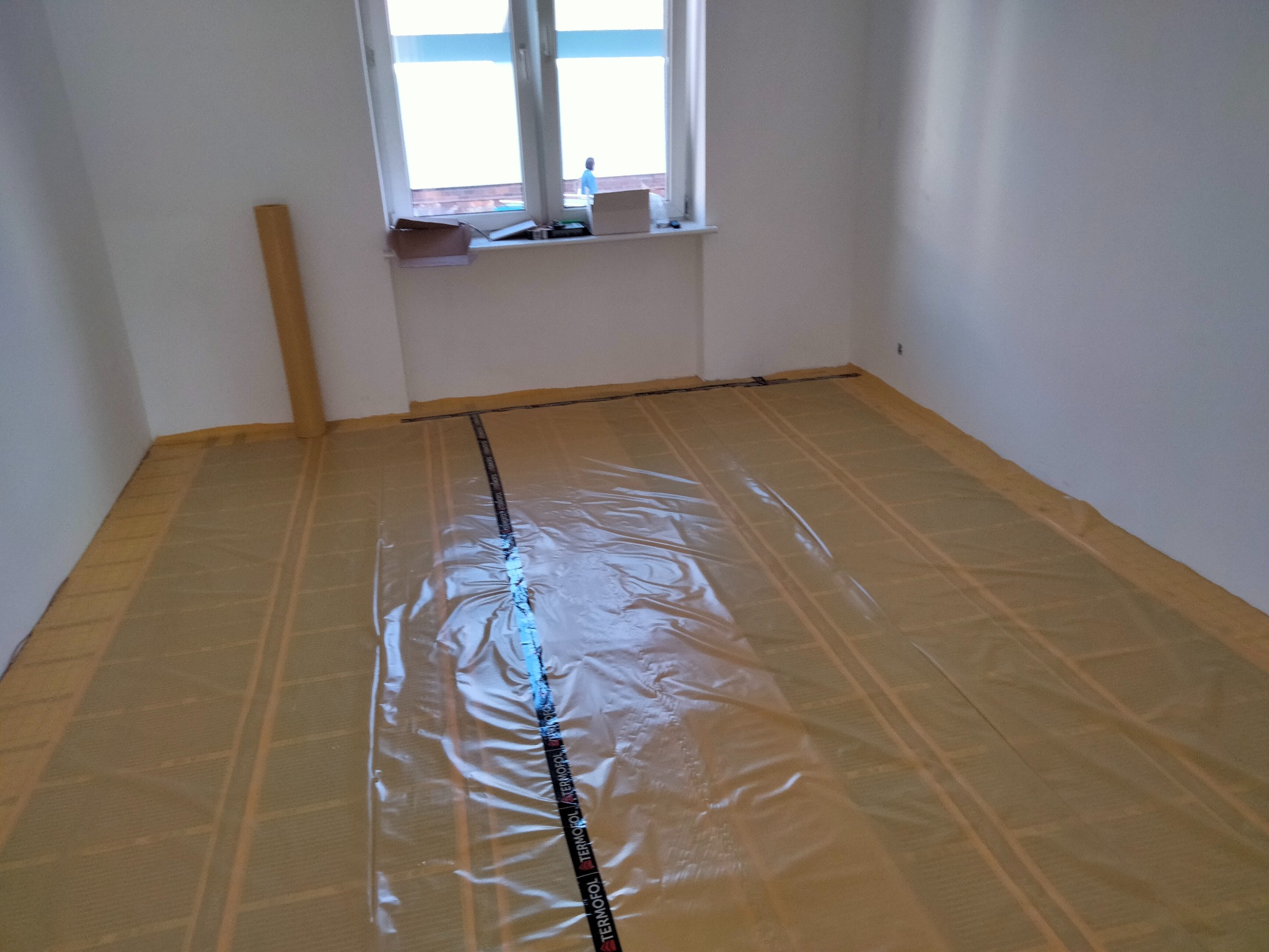 Przygotowanie podłoża pod ogrzewanie podłogowe: rozłożona folia izolacyjna Termofol na warstwie grzewczej w pustym, jasnym pomieszczeniu.