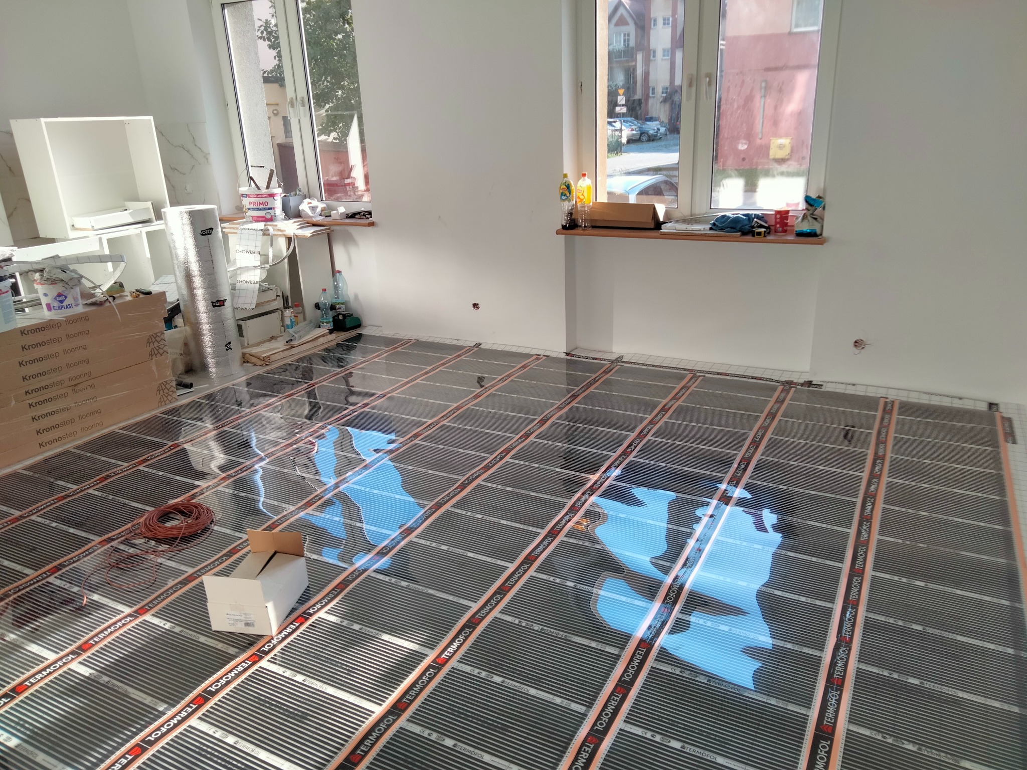 Montaż folii grzewczej Termofol na podłodze w jasnym pomieszczeniu, widoczne okna, kartony z panelami Kronostep i materiały budowlane.
