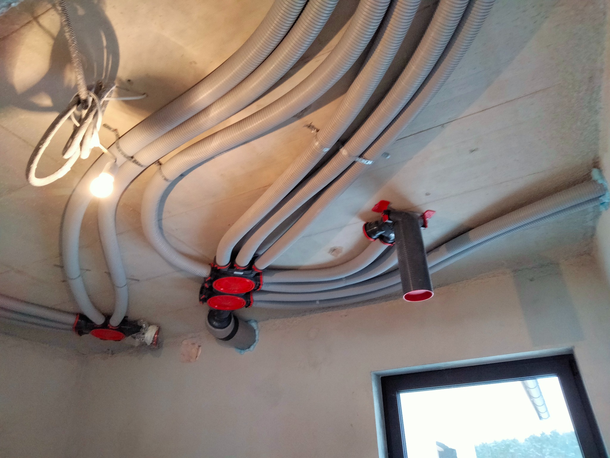 Szare, karbowane rury wentylacyjne prowadzone pod betonowym sufitem, połączone czerwonymi rozdzielaczami, z widocznym oknem w tle.