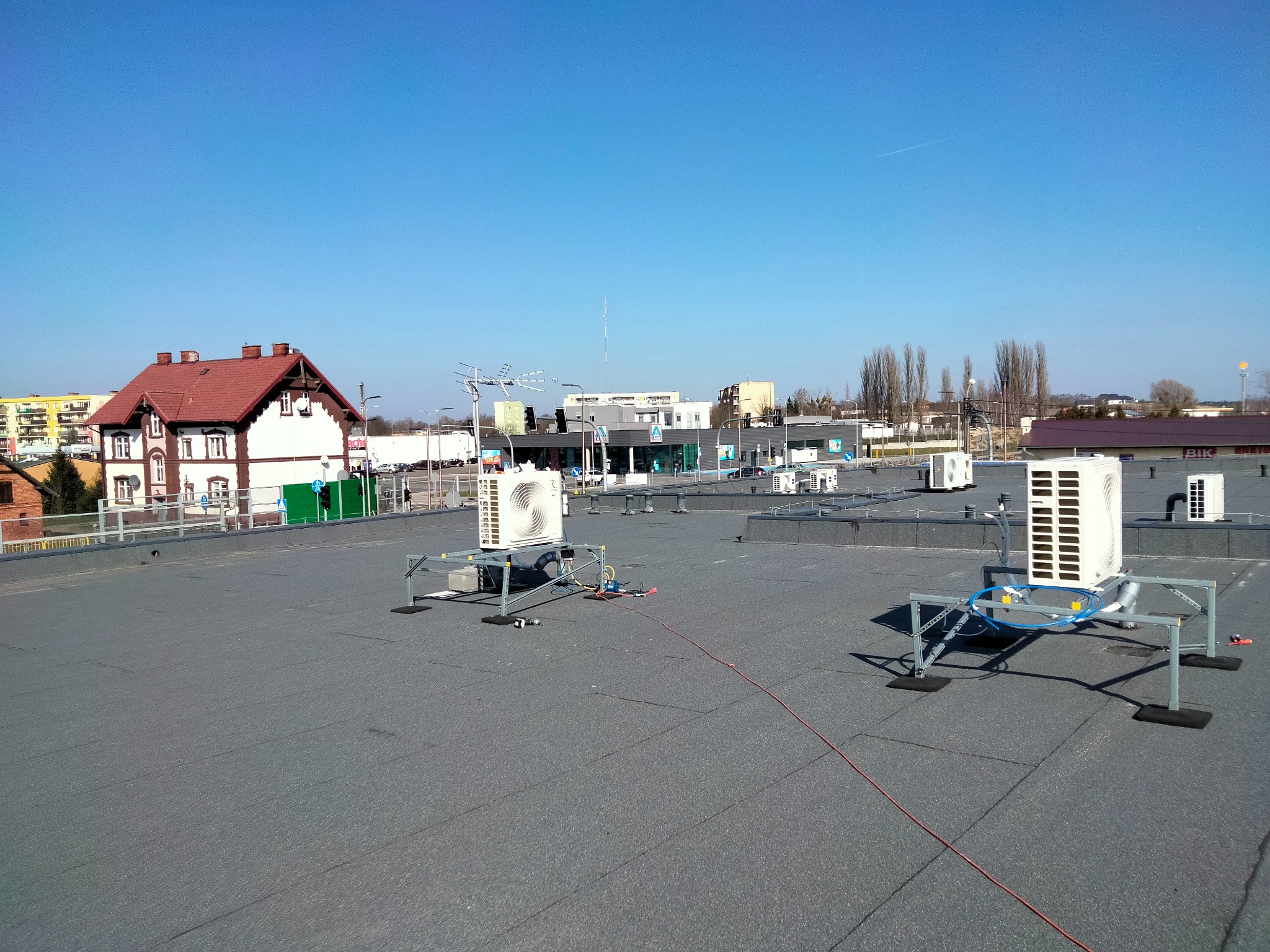 Widok z góry na płaski dach z kilkoma zewnętrznymi jednostkami klimatyzacji na metalowych konstrukcjach wsporczych, widoczne przewody i narzędzia sugerujące trwający montaż, w tle budynki miejskie...