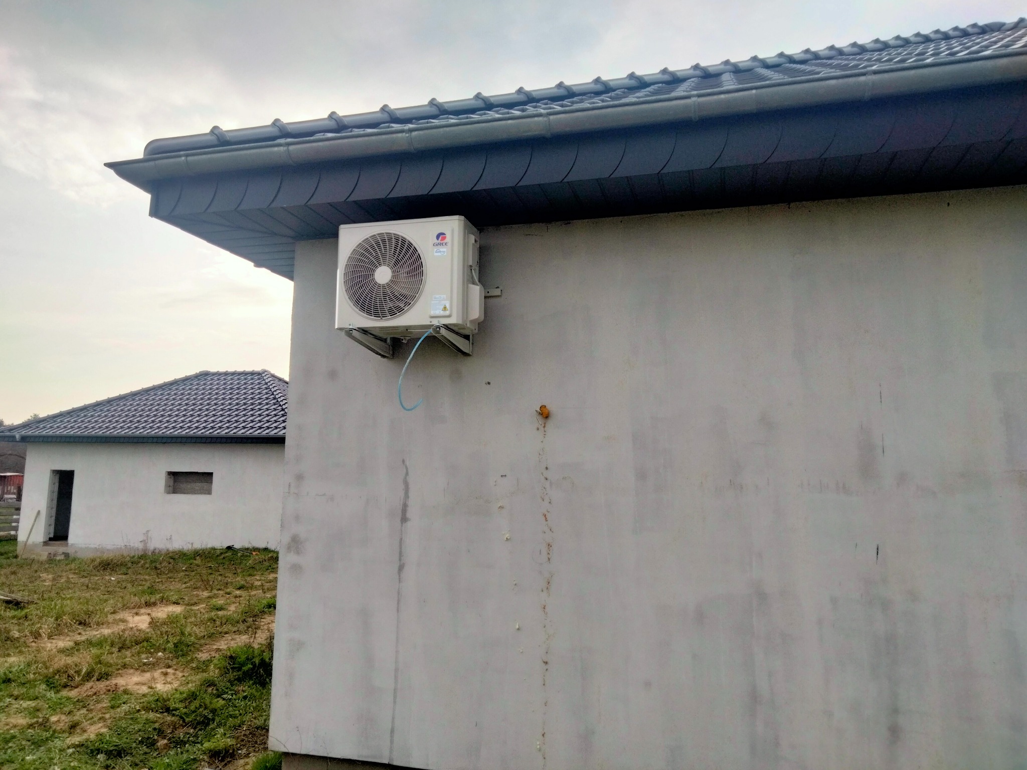 Zewnętrzna jednostka klimatyzacji marki Gree zamontowana na surowej, szarej ścianie budynku z ciemną, nowoczesną dachówką; widoczny fragment drugiego budynku w tle.