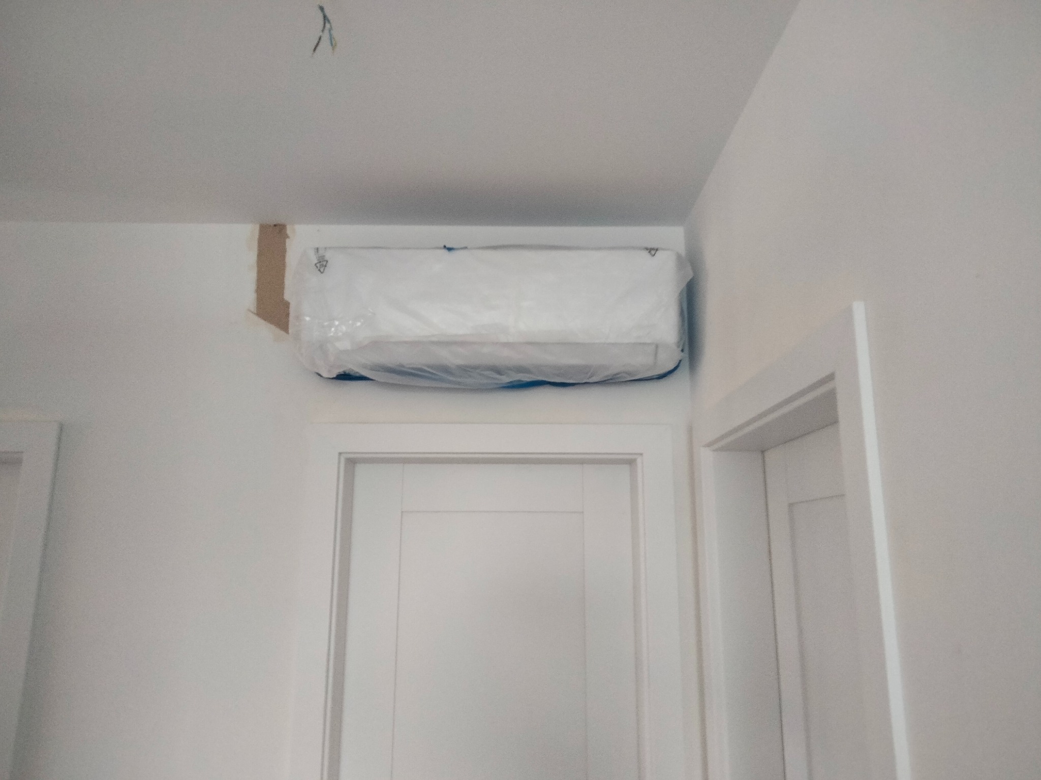 Nowy klimatyzator, jeszcze w folii ochronnej, zamontowany nad białymi drzwiami w białym pomieszczeniu, widoczne kable wystające ze ściany.