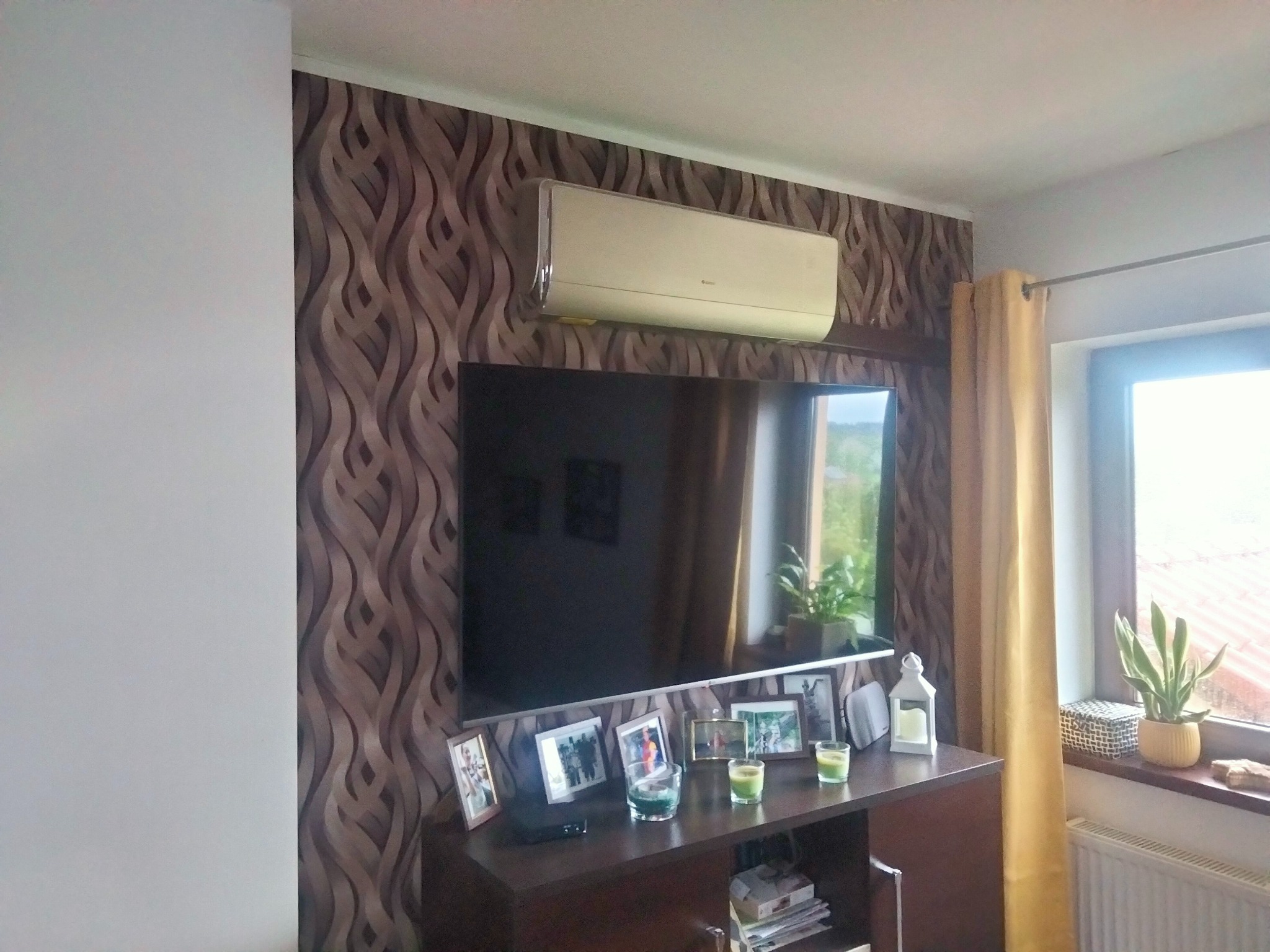 Klimatyzator zamontowany nad telewizorem w pokoju z wzorzystą tapetą, widoczne ramki ze zdjęciami i dekoracje na szafce RTV