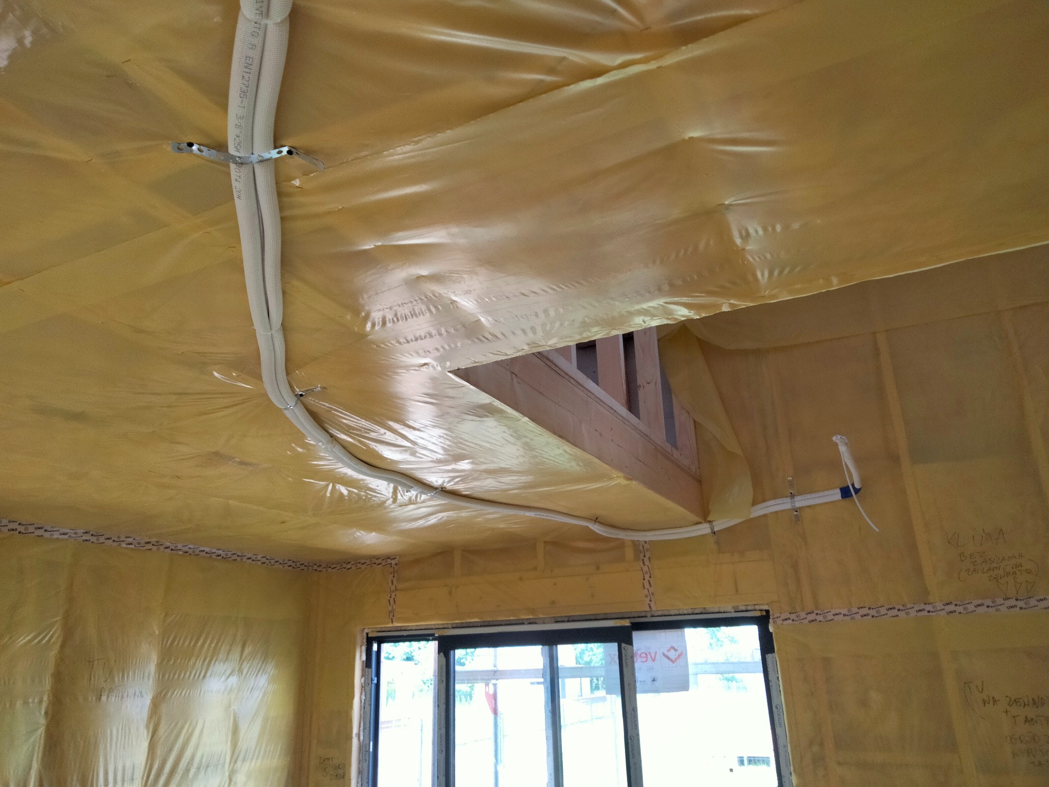 Montaż instalacji klimatyzacyjnej w nowym budynku: białe rury przymocowane do żółtej folii izolacyjnej na ścianach i suficie, widoczne drewniane elementy konstrukcyjne i okno balkonowe.