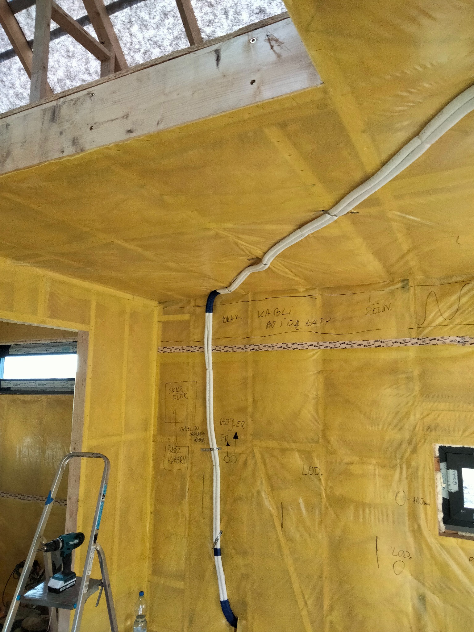 Wnętrze w trakcie instalacji klimatyzacji lub rekuperacji: widoczne przewody w izolacji, drabina z wiertarką, folia ochronna na ścianach z odręcznymi notatkami dotyczącymi instalacji elektrycznej...