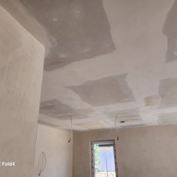 Remonty I wykonczenia Arkadiusz Gruca - Nierówna powierzchnia ścian i sufitu w trakcie prac wykończeniowych, widoczne ślady po szpachlowaniu i łączeniach płyt gipsowo-kartonowych, wystające przewody elektryczne, okno z widokiem na zieleń.