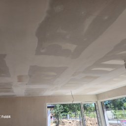 Remonty I wykonczenia Arkadiusz Gruca - Sufit w trakcie prac wykończeniowych, widoczne łączenia płyt gipsowo-kartonowych pokryte masą szpachlową, wystające przewody elektryczne, duże okna z widokiem na plac budowy.