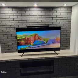 Telewizor zawieszony na ścianie z dekoracyjnej, szarej cegły, podświetlonej punktowymi lampami sufitowymi, z wnękami po bokach.