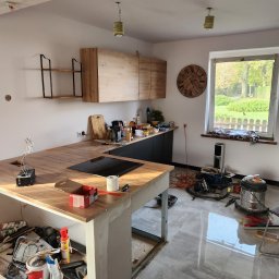 Remonty I wykonczenia Arkadiusz Gruca - Wnętrze kuchni w trakcie remontu w Wachowie: widoczne niedokończone zabudowy meblowe, surowy blat wyspy kuchennej z płytą indukcyjną, oraz narzędzia i materiały budowlane rozłożone na podłodze...