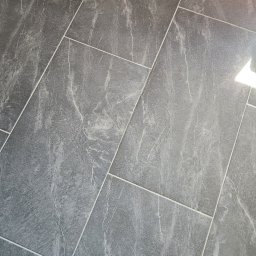 Remonty I wykonczenia Arkadiusz Gruca - Szare płytki podłogowe imitujące marmur, ułożone ukośnie, widoczne fugi i odbicie światła.