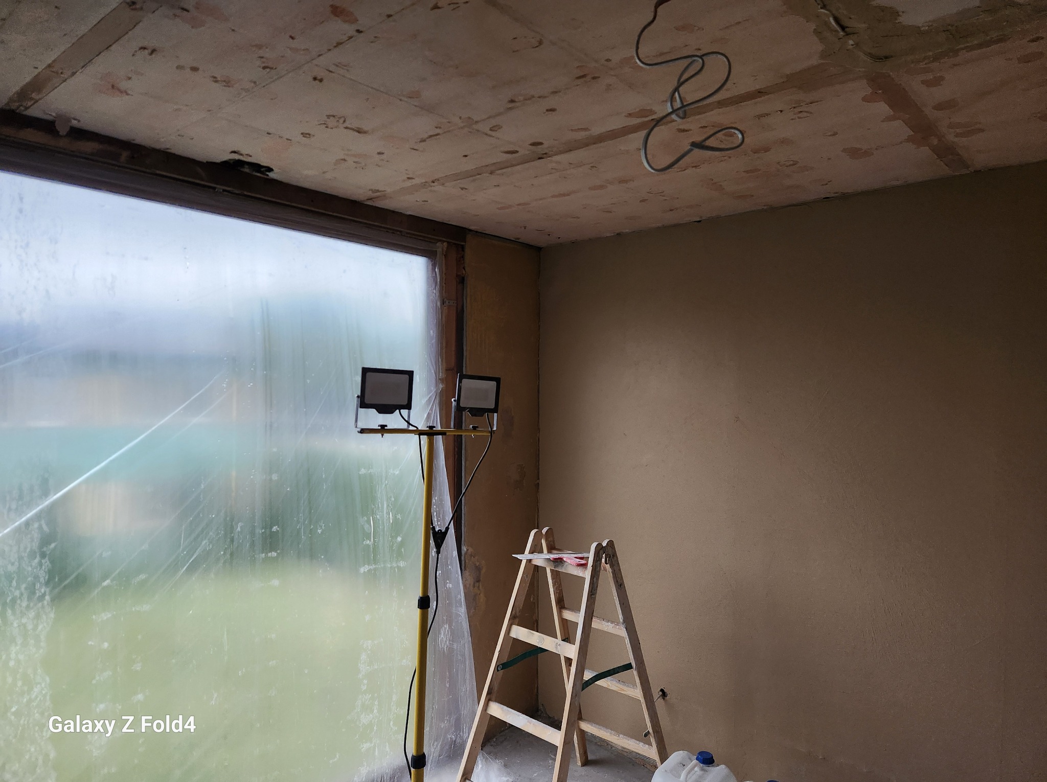 Wnętrze w trakcie prac przygotowawczych: ściany pokryte świeżym tynkiem, okno zabezpieczone folią, ustawiona drabina i lampa budowlana, widoczne przewody elektryczne na suficie.