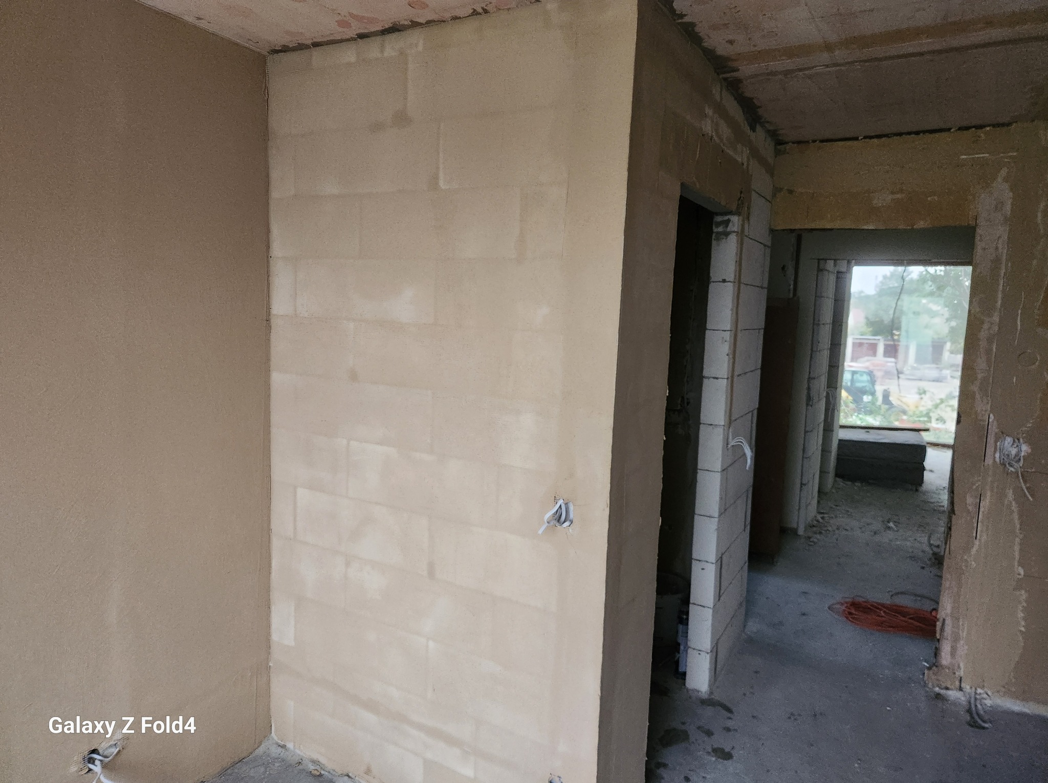 Wnętrze w trakcie szpachlowania: ściany z widocznymi blokami i gładzią, wystające kable elektryczne, surowy betonowy sufit, widok na kolejne pomieszczenia w tle.