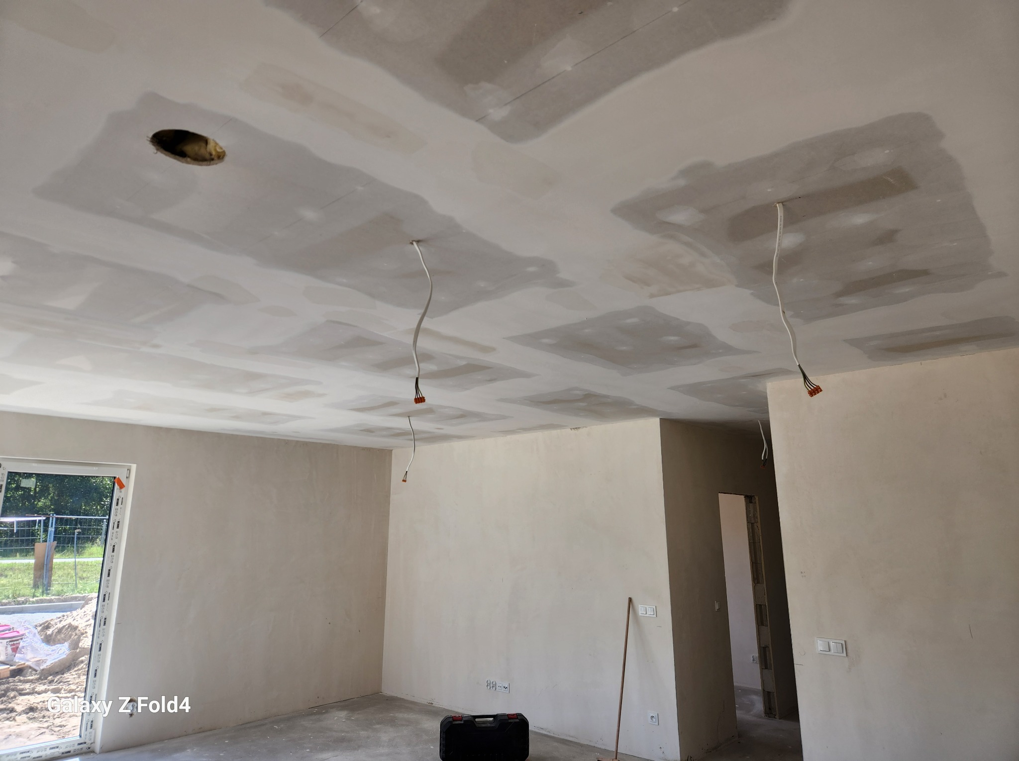 Wnętrze w trakcie prac wykończeniowych: sufit z płyt gipsowo-kartonowych z widocznymi łączeniami wymagającymi szpachlowania, wystające przewody elektryczne, ściany po wstępnym przygotowaniu, czarna...