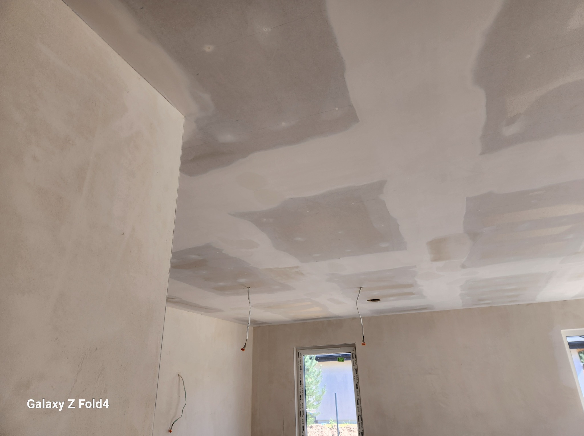 Nierówna powierzchnia ścian i sufitu w trakcie prac wykończeniowych, widoczne ślady po szpachlowaniu i łączeniach płyt gipsowo-kartonowych, wystające przewody elektryczne, okno z widokiem na zieleń.