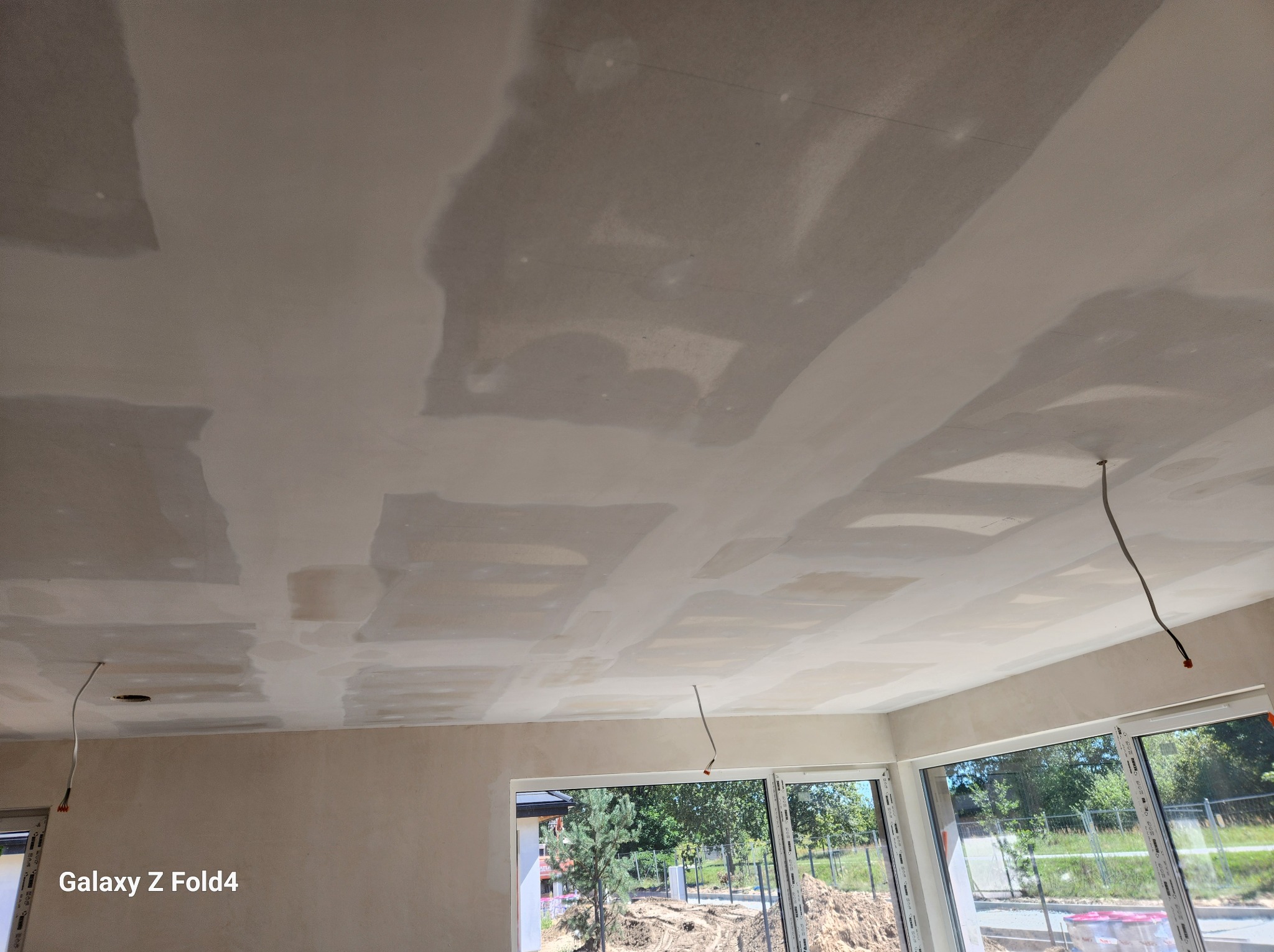 Sufit w trakcie prac wykończeniowych, widoczne łączenia płyt gipsowo-kartonowych pokryte masą szpachlową, wystające przewody elektryczne, duże okna z widokiem na plac budowy.