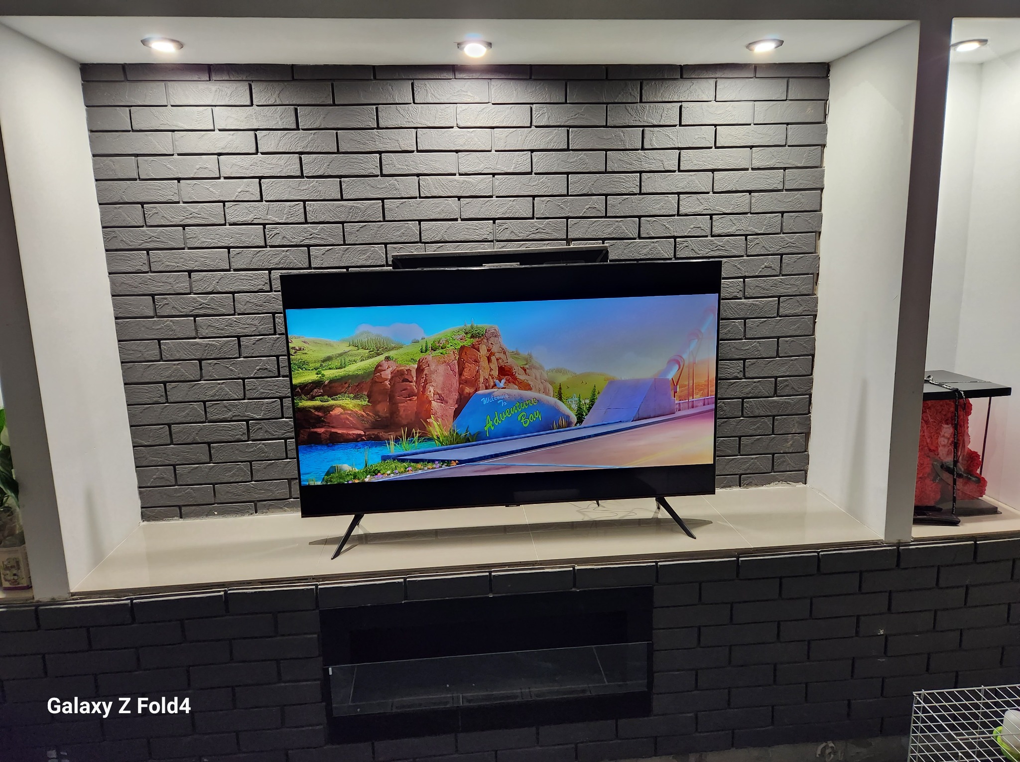 Telewizor zawieszony na ścianie z dekoracyjnej, szarej cegły, podświetlonej punktowymi lampami sufitowymi, z wnękami po bokach.