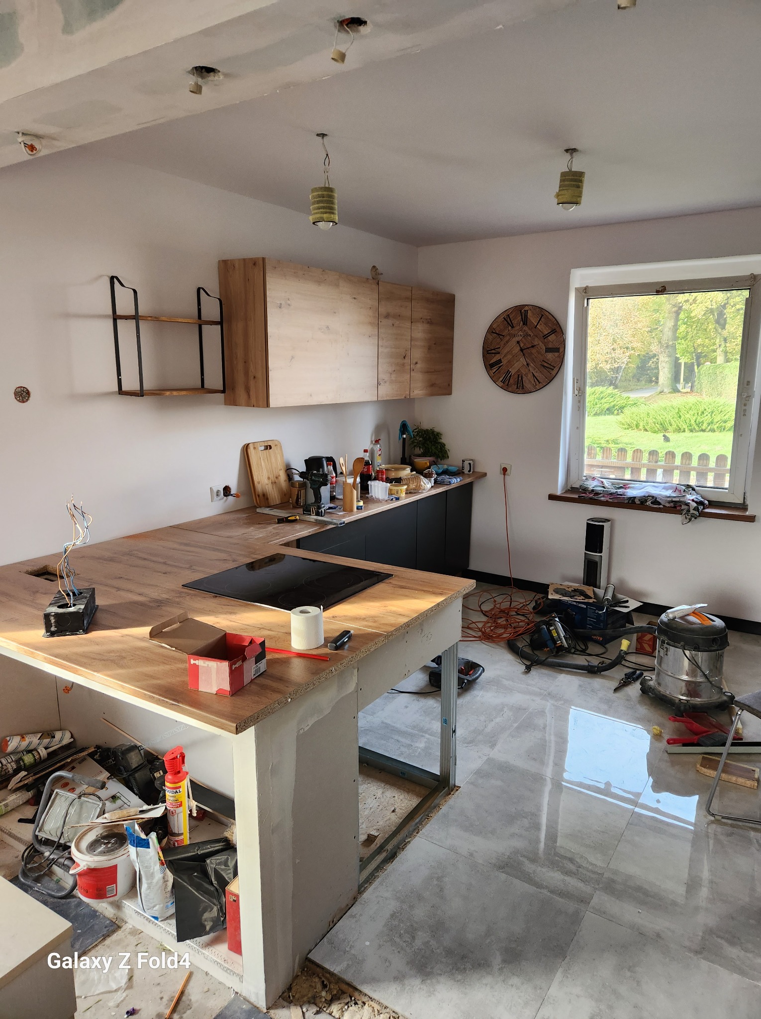 Wnętrze kuchni w trakcie remontu w Wachowie: widoczne niedokończone zabudowy meblowe, surowy blat wyspy kuchennej z płytą indukcyjną, oraz narzędzia i materiały budowlane rozłożone na podłodze...