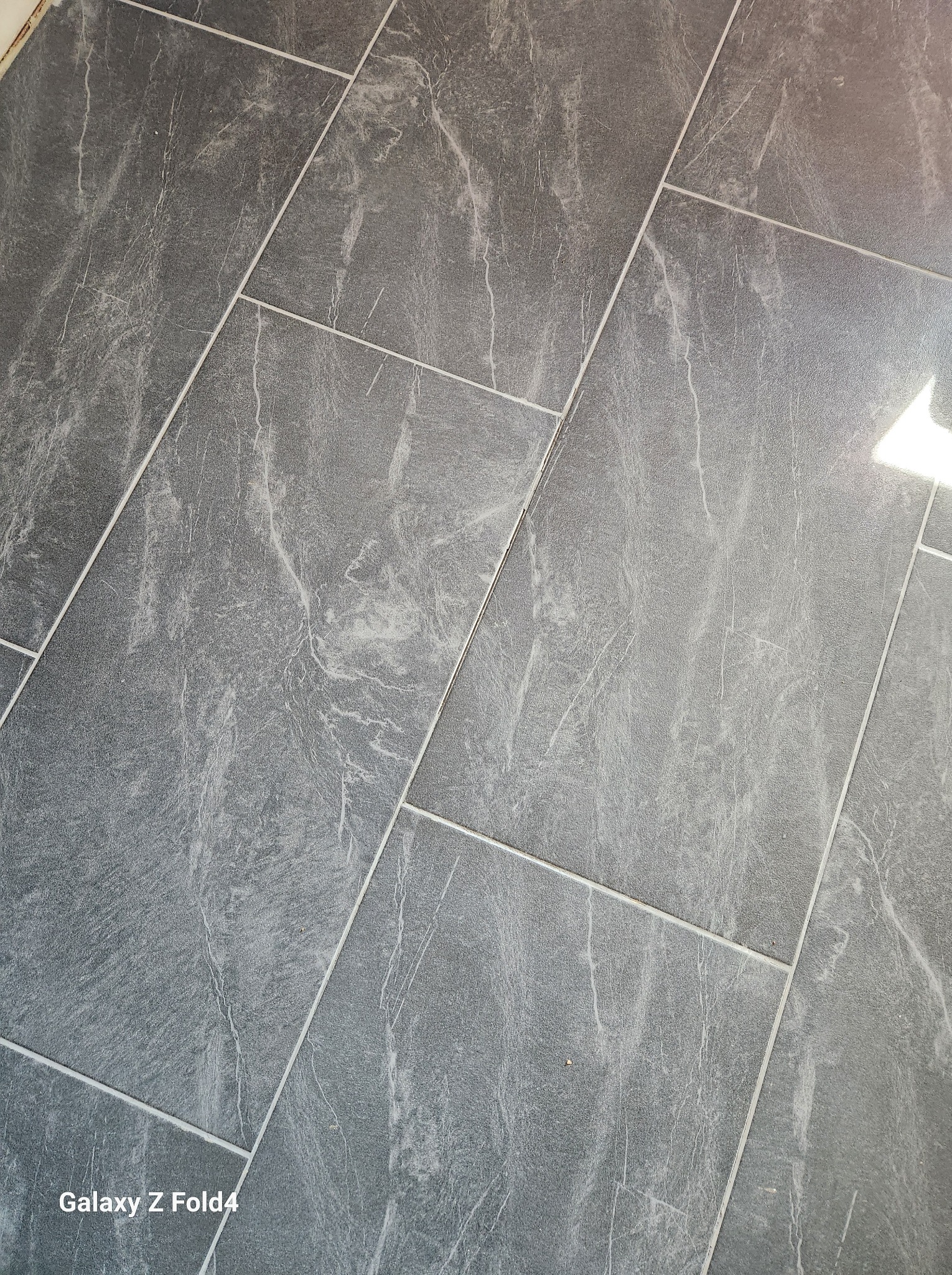 Szare płytki podłogowe imitujące marmur, ułożone ukośnie, widoczne fugi i odbicie światła.