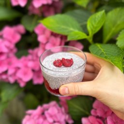 SS GASTRO SPÓŁKA Z OGRANICZONĄ ODPOWIEDZIALNOŚCIĄ - Deser chia z malinami w szklance, trzymany w dłoni na tle różowych hortensji. Zdjęcie z bliska, oświetlone naturalnym światłem, eksponuje teksturę deseru.