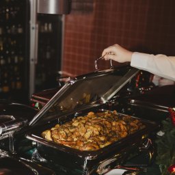 SS GASTRO SPÓŁKA Z OGRANICZONĄ ODPOWIEDZIALNOŚCIĄ - Podgrzewacz bufetowy ze złotymi ziemniakami, otwarty przez dłoń w białej koszuli. Dekoracja świąteczna, zielony obrus. Elegancki catering.