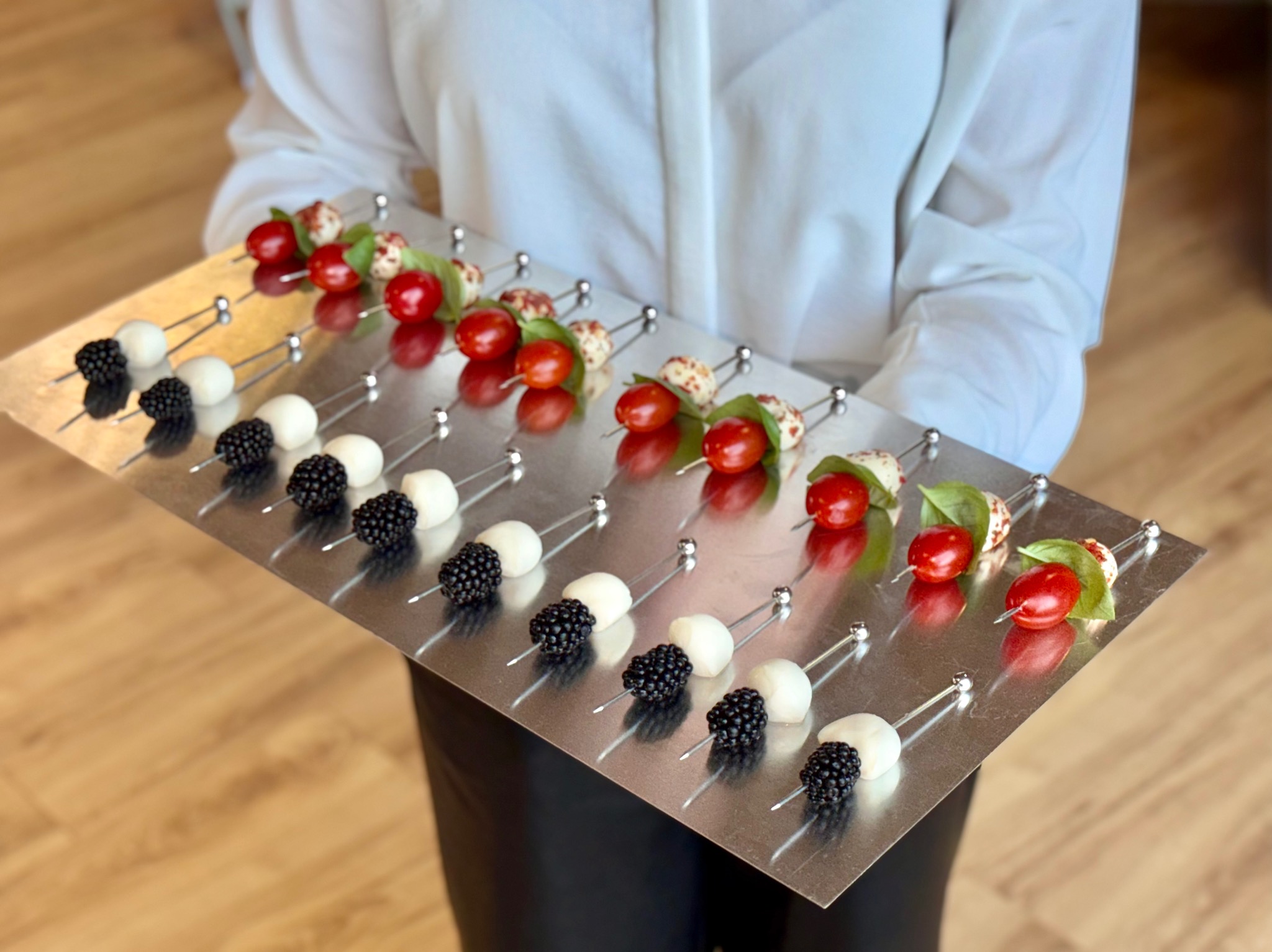 Osoba prezentuje metalową tacę z apetycznymi koreczkami: mozzarella z jeżyną i pomidorki cherry z bazylią, idealne na elegancki catering.