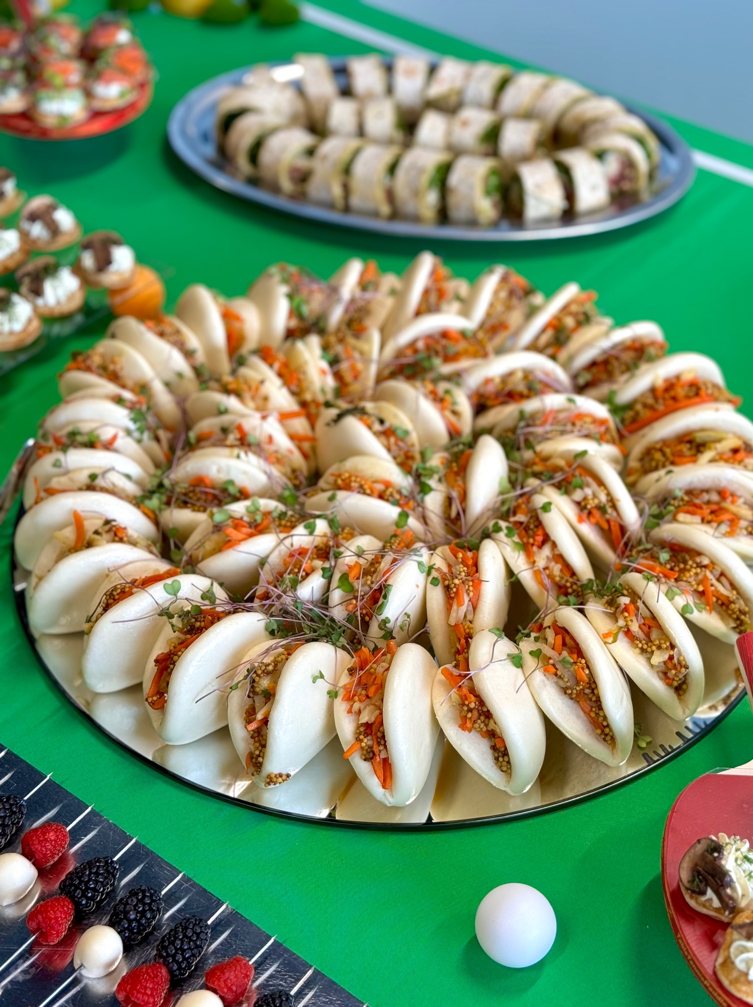 Elegancko ułożone bułeczki bao z warzywnym farszem na lustrzanej tacy, otoczone innymi przekąskami na zielonym tle. Dekoracja i catering wizualnie zachęcają do spożycia.