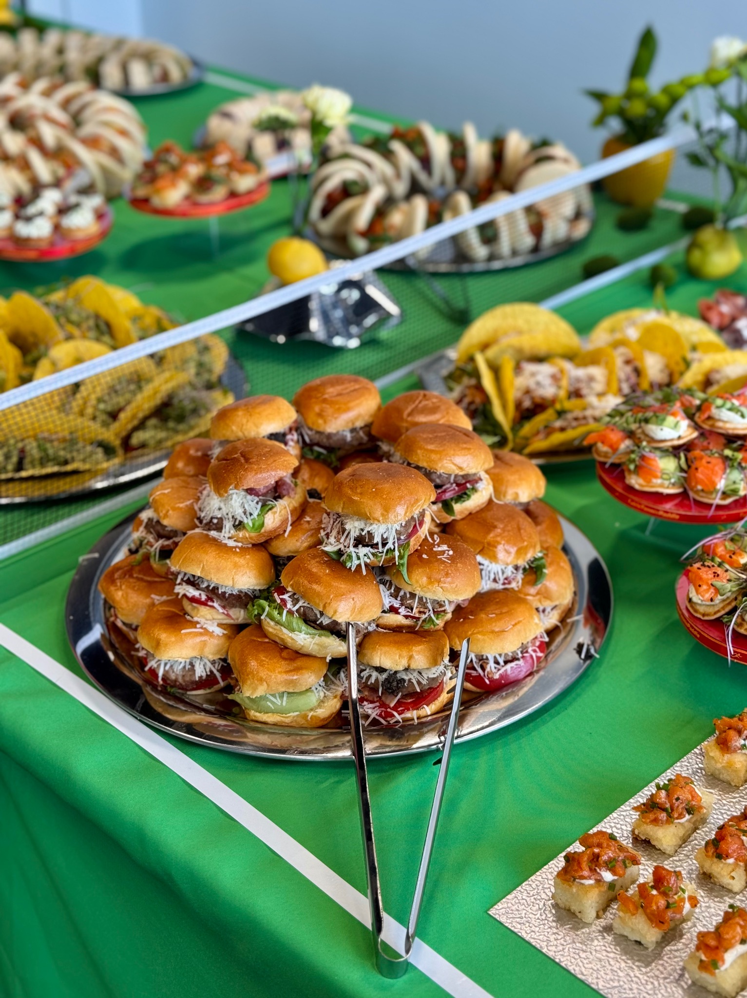 Bogato zastawiony stół cateringowy z mini burgerami, tacos, kanapeczkami i innymi przekąskami na zielonym obrusie. Widoczne szczypce do nakładania.