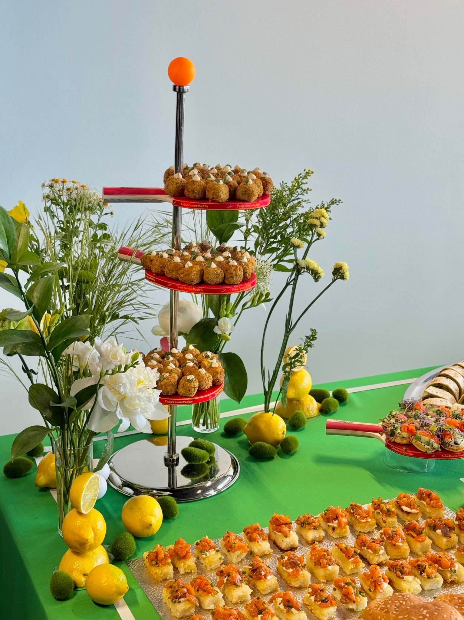 Stół z zielonym obrusem, dekoracje z cytryn i mchu, trzypoziomowy pater z przekąskami. Dekoracje kwiatowe w wazonach. Elementy nawiązujące do tenisa stołowego.
