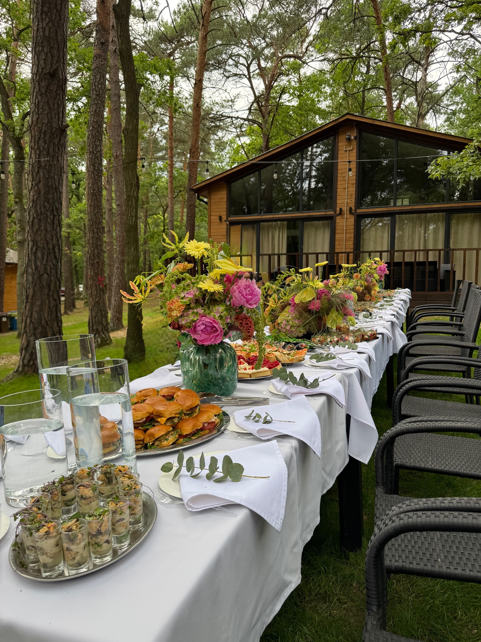 Stół cateringowy na zewnątrz z burgerami, przekąskami w szklankach i kwiatami. W tle drewniany budynek i las.