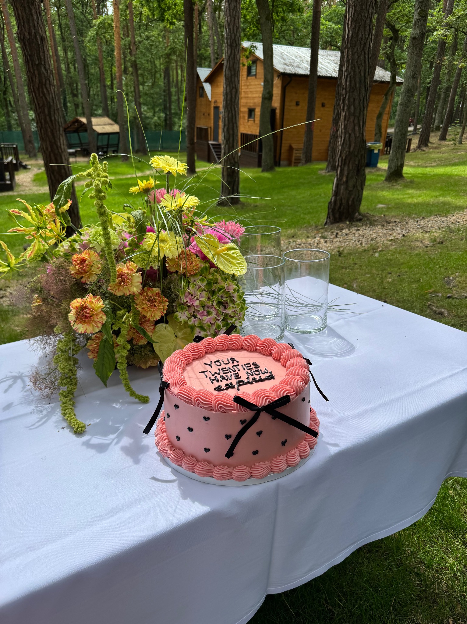 Różowy tort z czarnymi wstążkami i napisem, bukiet kwiatów i szklanki na białym obrusie, drewniany budynek w tle, plenerowa uroczystość.