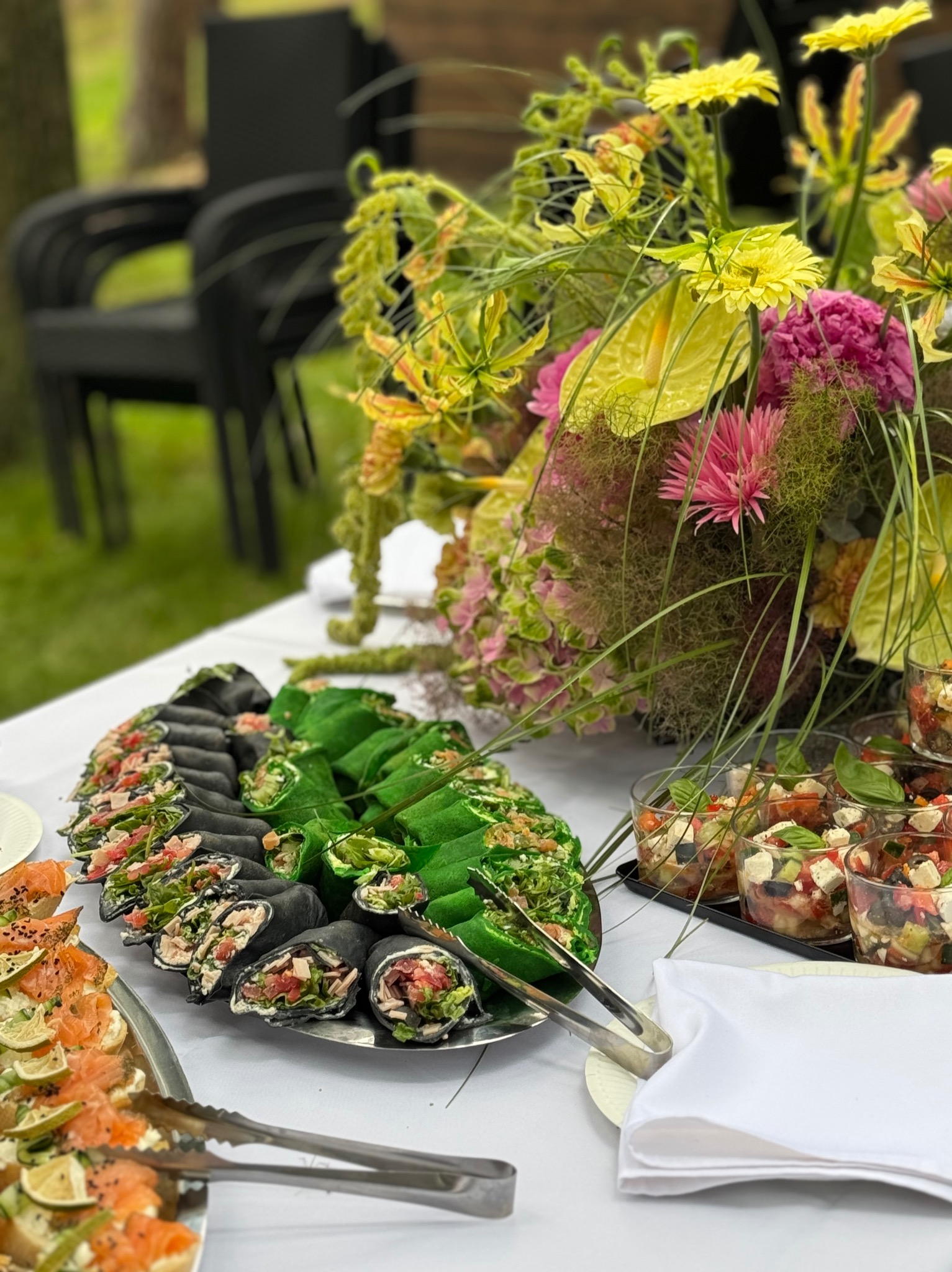 Stół z kolorowymi roladkami, sałatkami w szklankach i kwiatową dekoracją. Czarno-zielone wrap-y, przekąski z łososiem i szczypcami, elegancki catering.