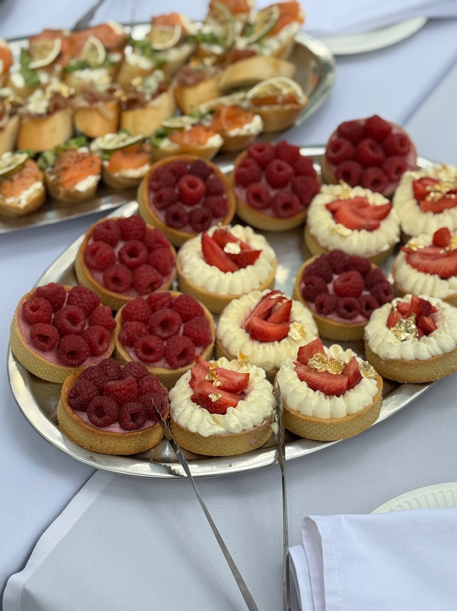 Tarty z malinami i truskawkami, udekorowane złotem, na srebrnej tacy obok kanapek z łososiem, elegancki catering.