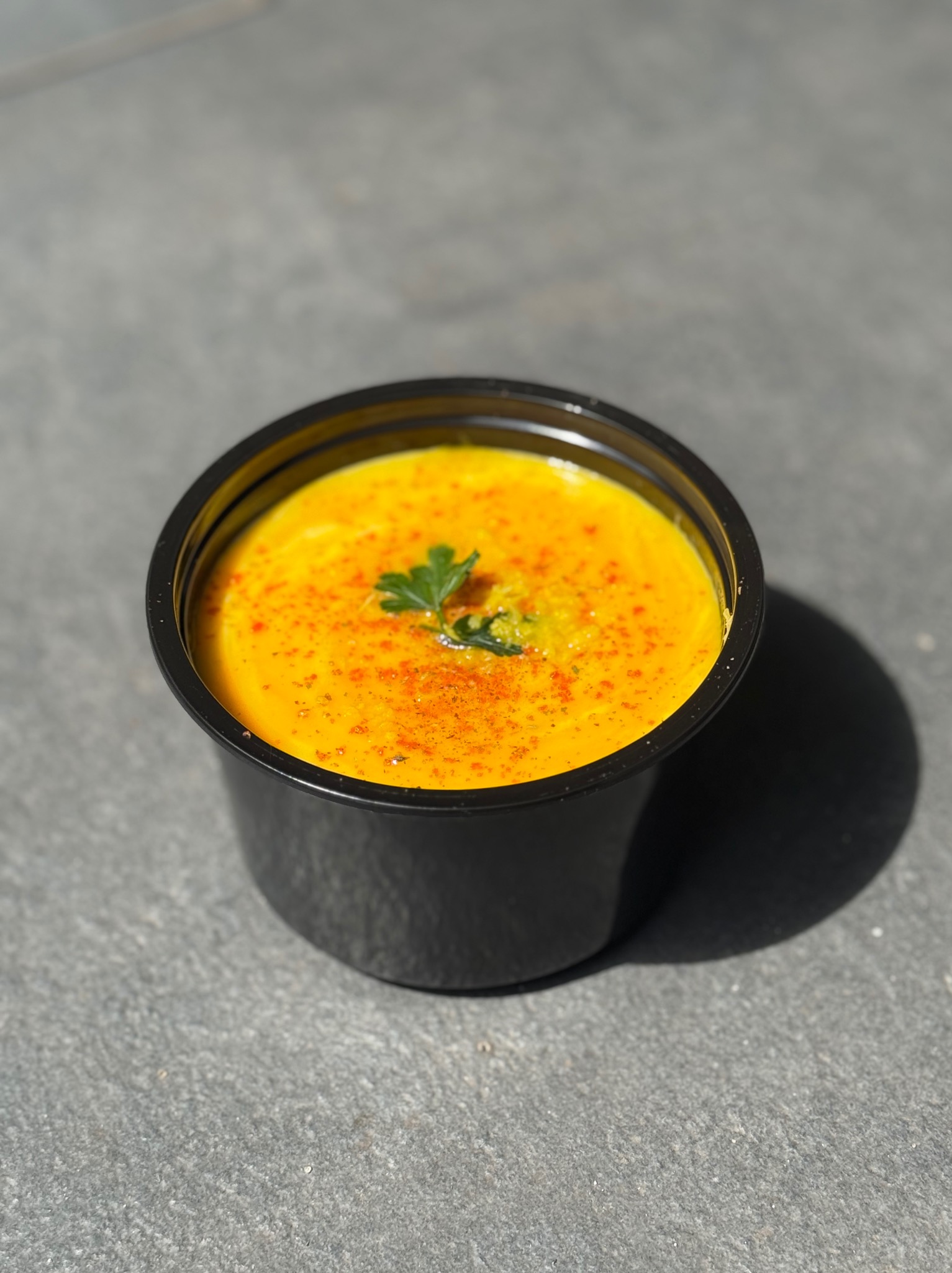 Porcja kremowej zupy dyniowej w czarnym pojemniku, posypana papryką i udekorowana zieloną pietruszką, na szarym tle.