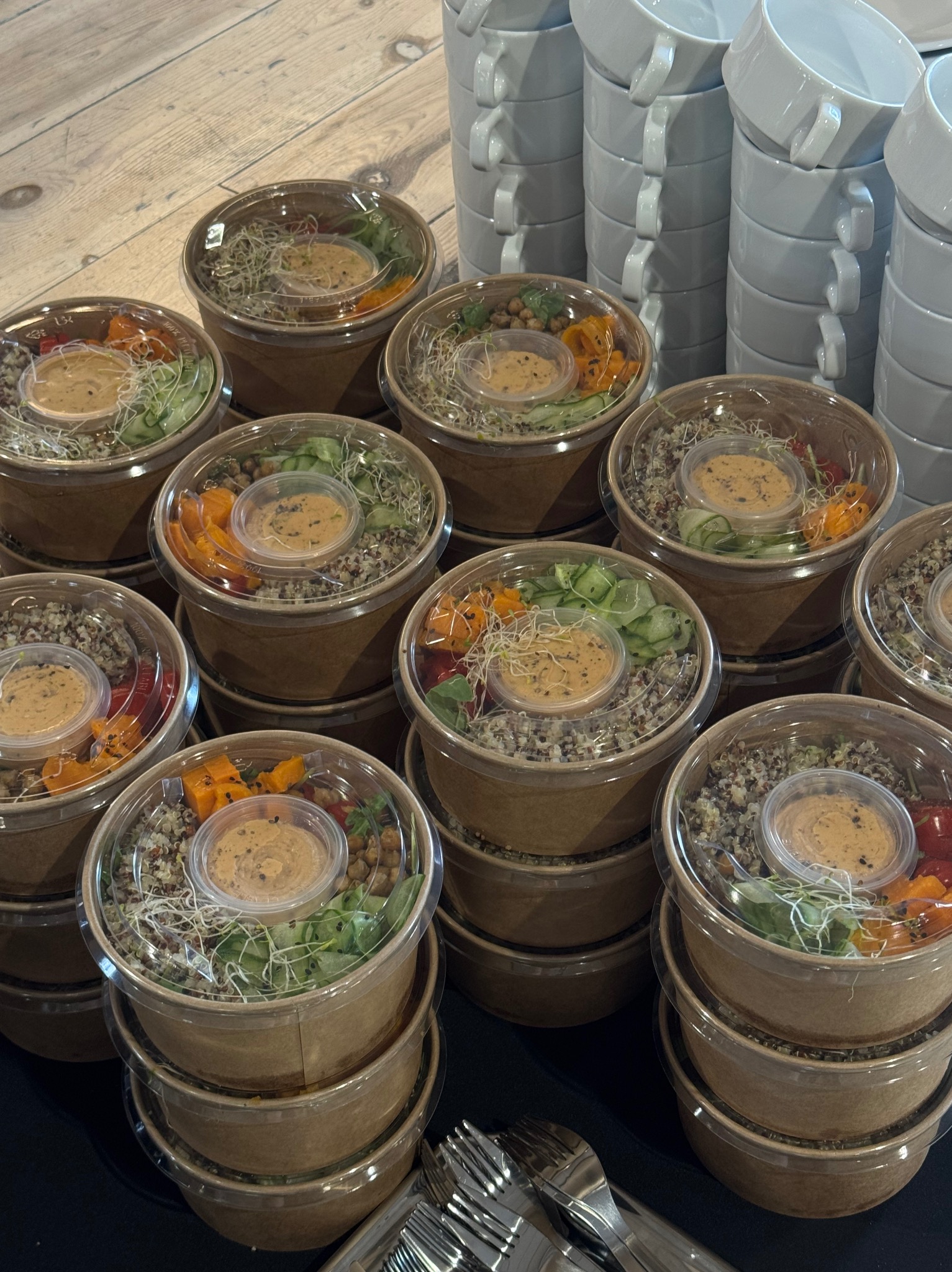 Zdrowe, gotowe dania w papierowych miskach z przezroczystymi pokrywkami, ułożone w stosy obok stosu białych filiżanek i sztućców, gotowe do serwowania.