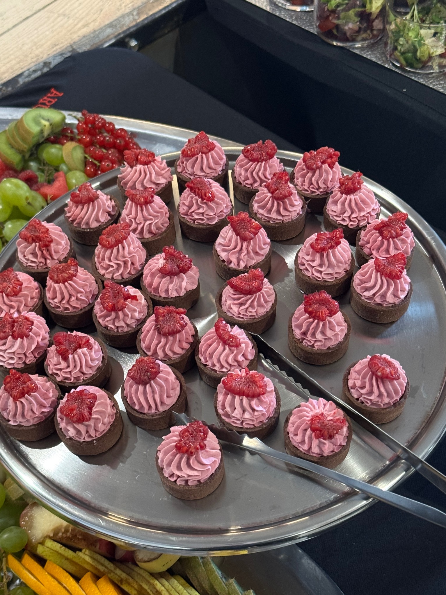 Srebrna taca z mini tartaletkami z różowym kremem i malinami, otoczona owocami (kiwi, winogrona, pomarańcze). Szczypce do nakładania. Elegancki catering.