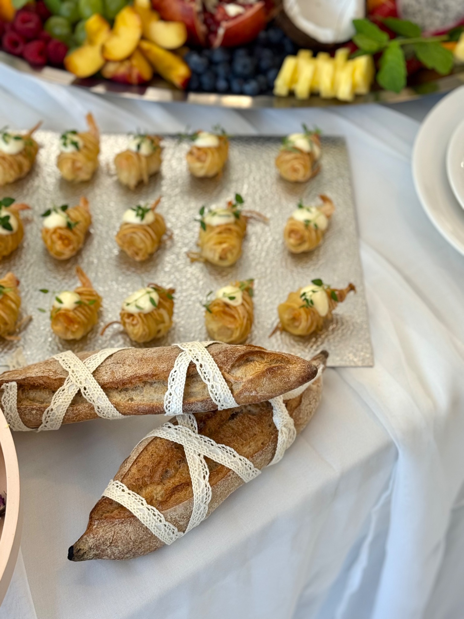 Elegancko podane przekąski na srebrnej tacy, z owocami w tle i bagietkami ozdobionymi koronką, tworzące wyrafinowaną kompozycję cateringową.