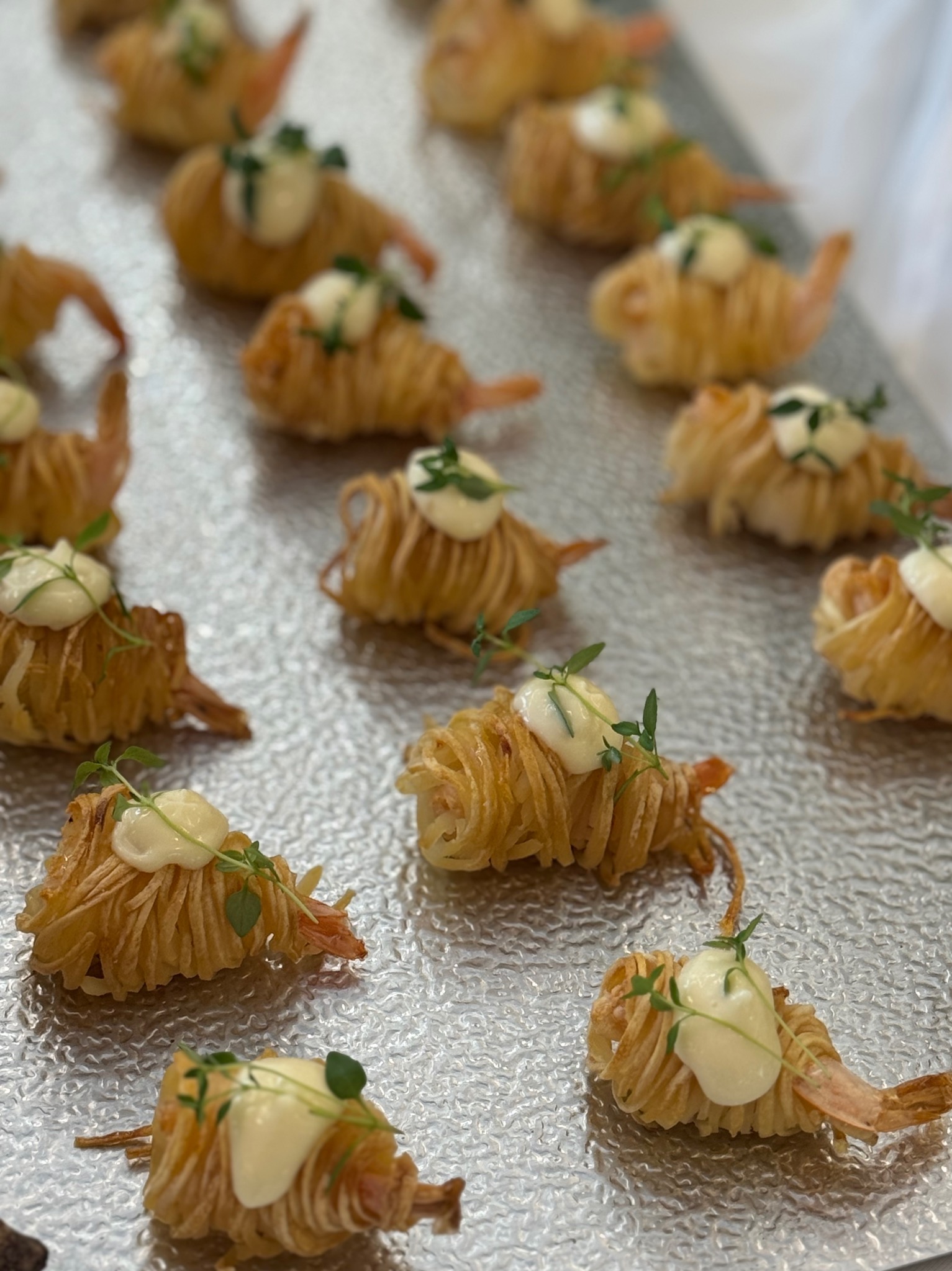 Ekskluzywne, zawijane krewetki z sosem i ziołami na srebrnej tacy, prezentowane podczas cateringu. Nietypowa forma podania, dekoracja z świeżych ziół.