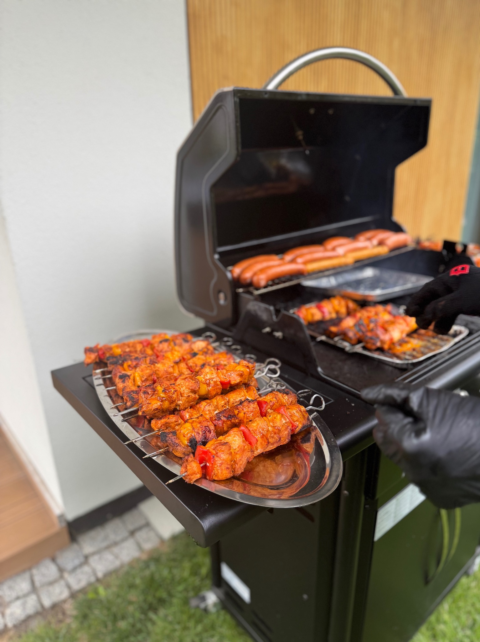 Przygotowane szaszłyki i kiełbaski na grillu gazowym, ręce w czarnych rękawicach obracają potrawy. Grill stoi na trawie, w tle jasna ściana.